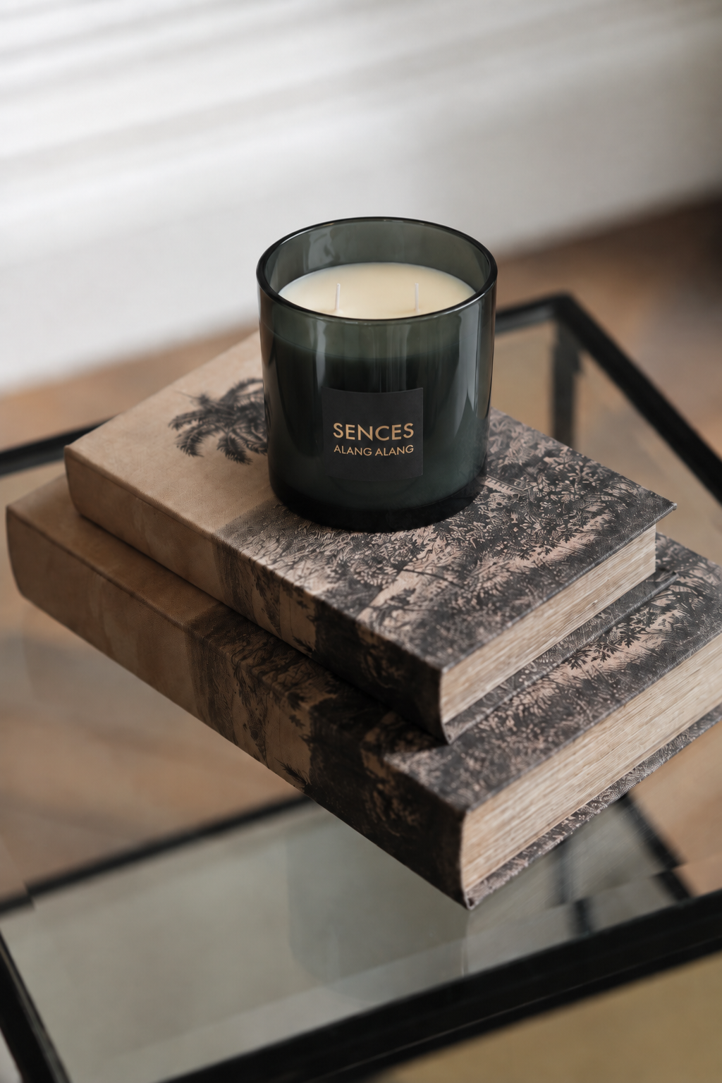 A dark smoky glass candle jar with cream-colored wax sits on top of two stacked vintage books on a glass table. The candle features a black label with the brand name 'SENCES' and the scent 'ALMO AOFO'.