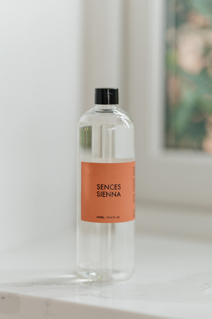 A clear glass bottle of Sences Sienna room spray with a black cap and a peach-colored label, standing on a white marble surface with a softly blurred window background.