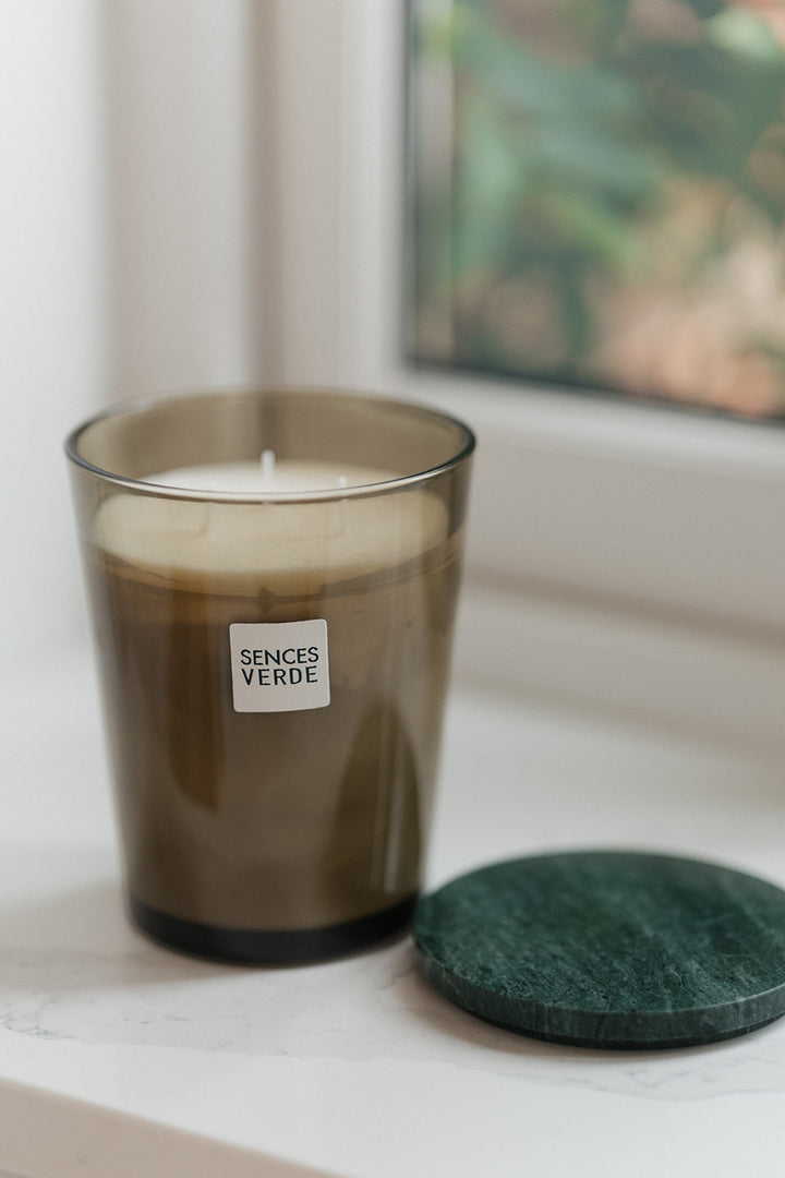 A Sentes Verde scented candle in a translucent dark amber glass jar sits on a white marble surface next to a dark green stone coaster, with a softly blurred window background.