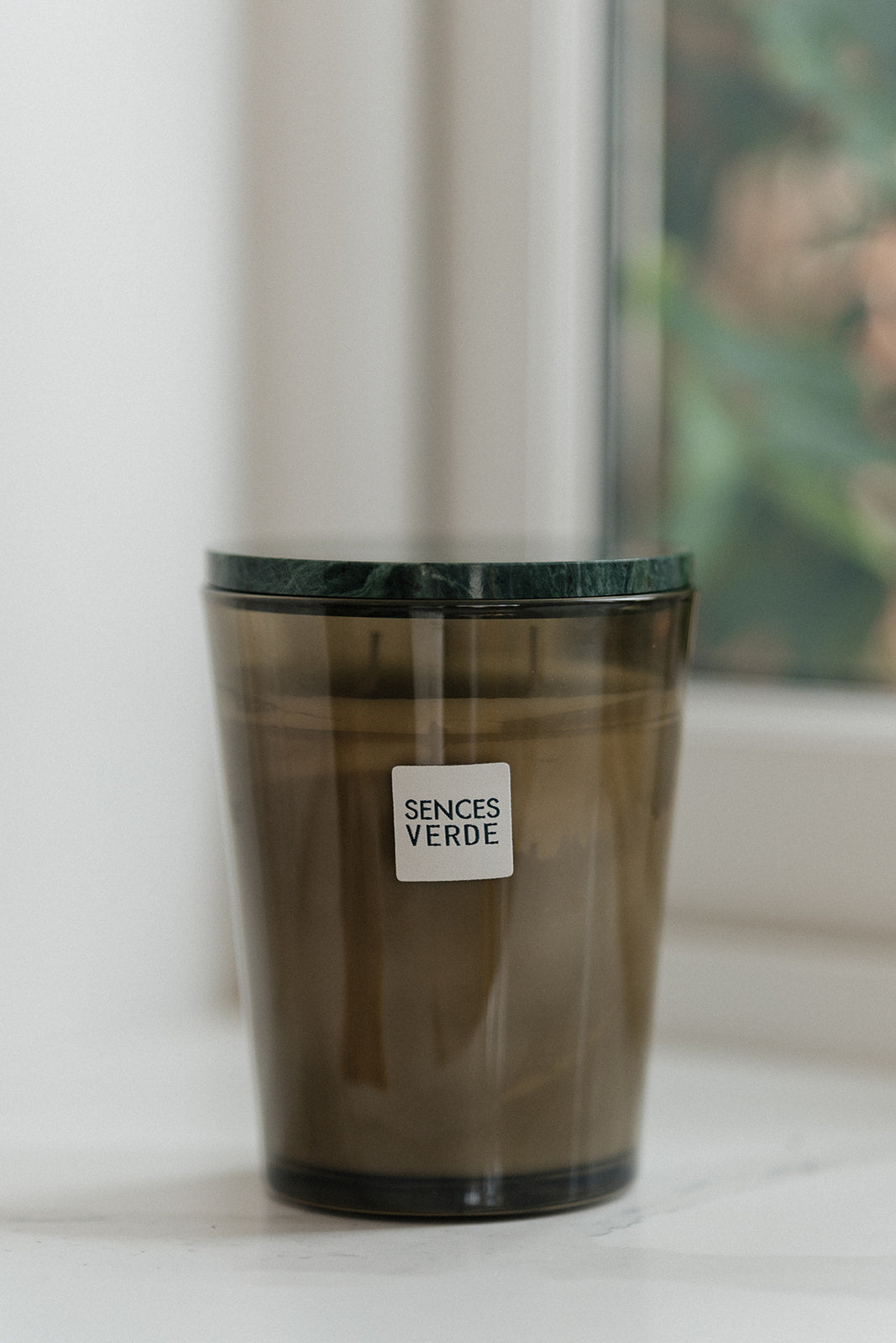 A dark amber glass jar candle with a black lid sits on a white windowsill. A small white label on the front reads &