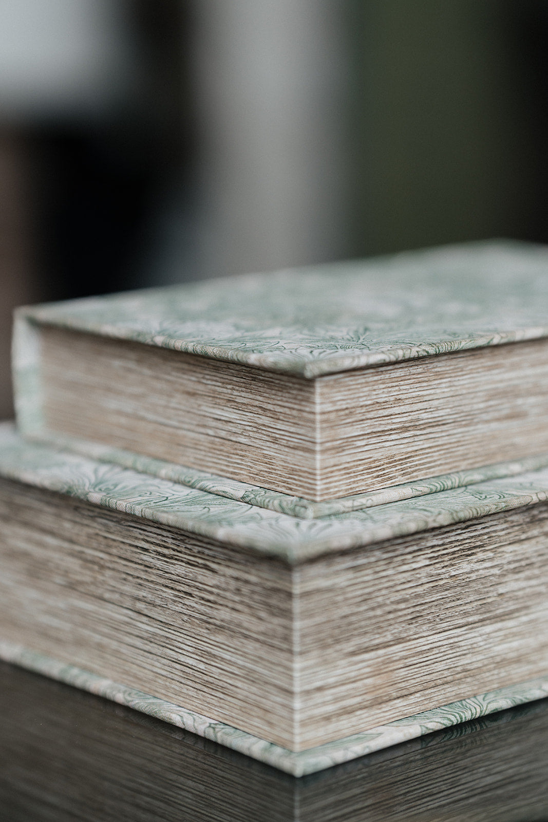 Two stacked hardcover books with textured sage green and beige patterned covers and thick, ribbed beige pages, resting on a dark reflective surface with a blurred background.
