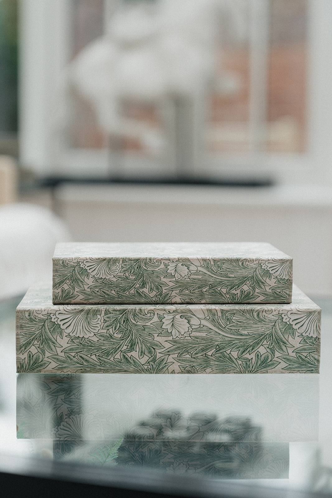 Two rectangular boxes stacked on top of each other, featuring a green botanical pattern on a light beige background, displayed on a reflective glass surface in a softly blurred indoor setting.