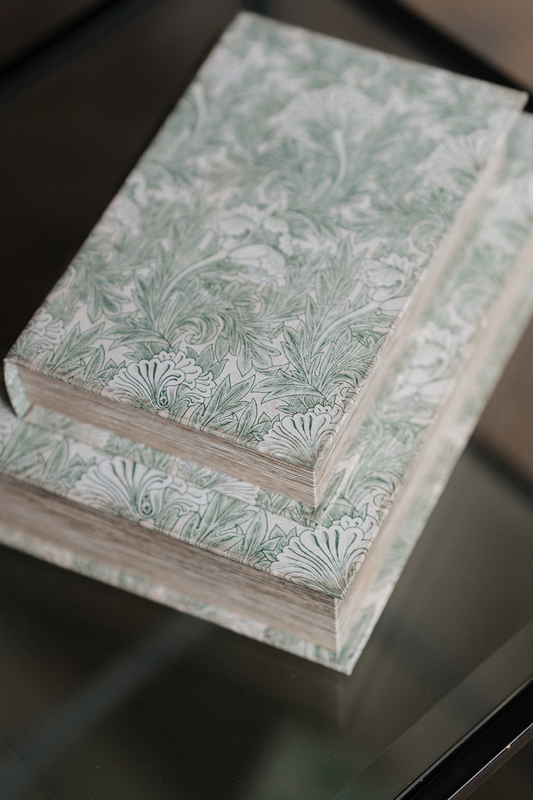 Two hardcover books stacked on a reflective dark glass surface, featuring a light sage green and white botanical pattern on the covers.