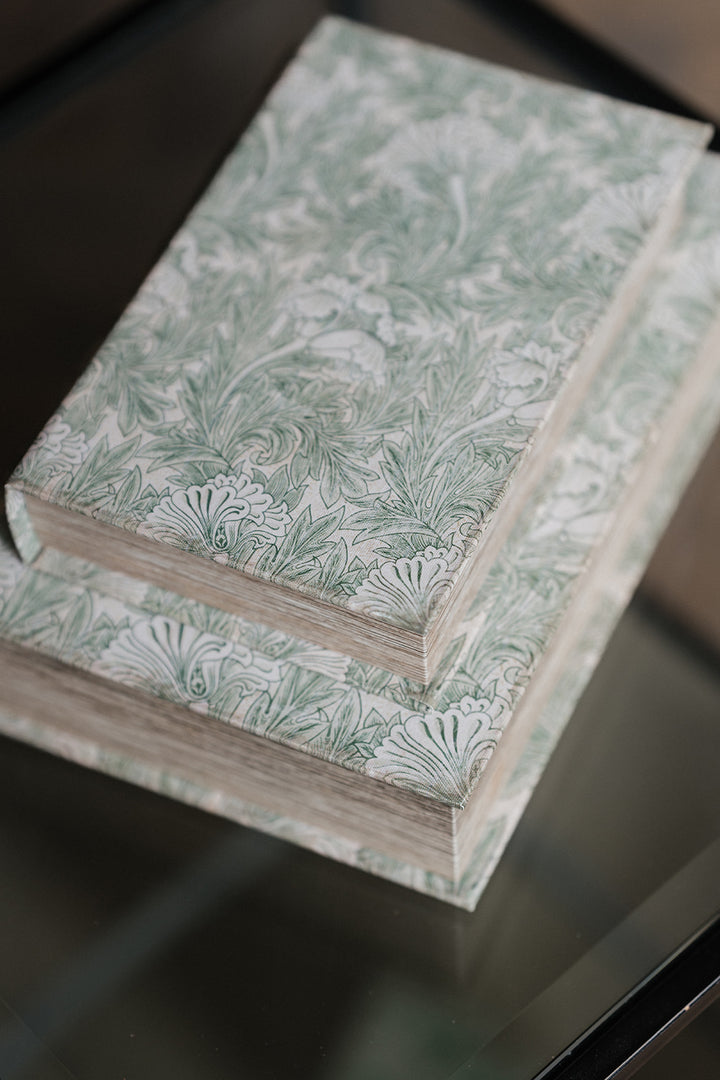Two hardcover books stacked on a reflective dark glass surface, featuring a light sage green and white botanical pattern on the covers.