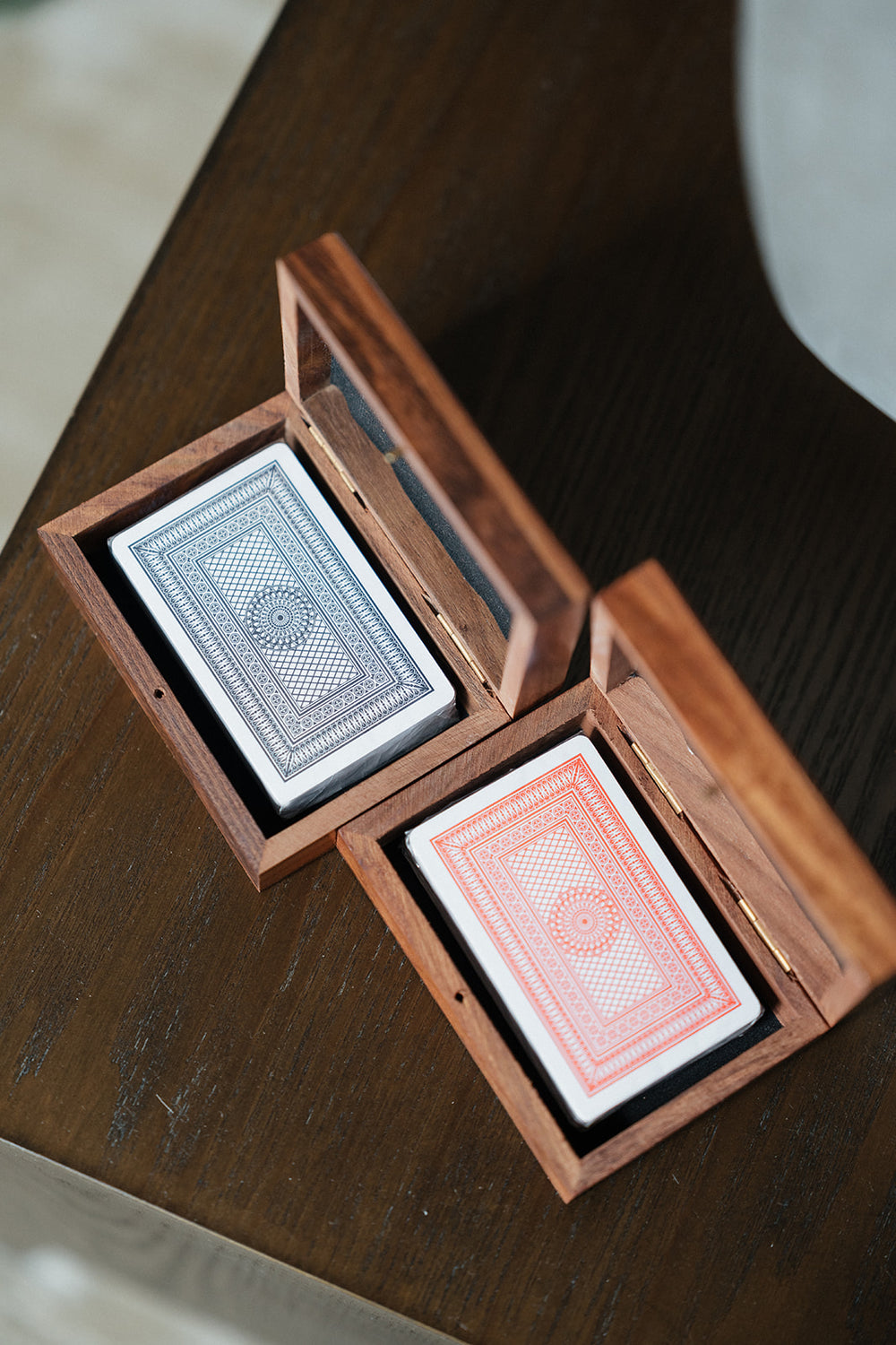 Two wooden deck boxes, one open to reveal a blue-gray patterned deck and the other open to reveal a coral-red patterned deck, resting on a dark wooden table.