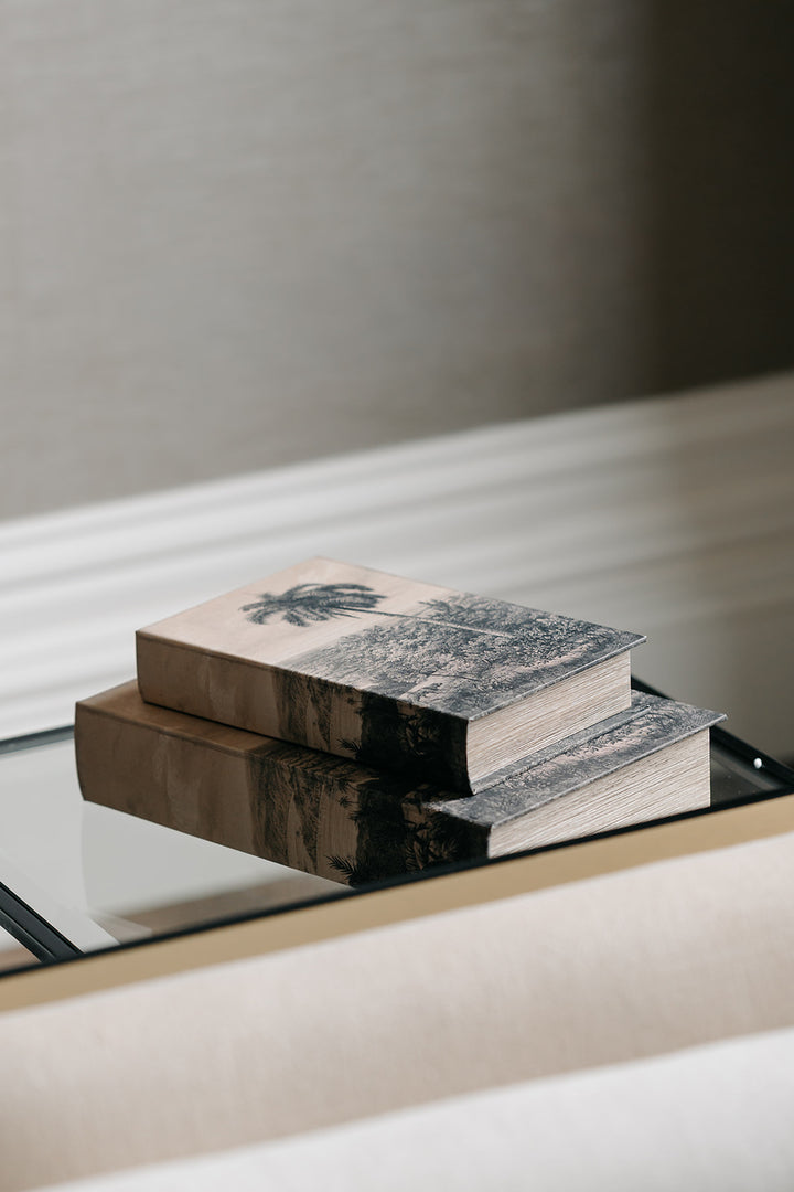 Two decorative books stacked on a glass tray, featuring a black and white landscape design on the top book and a warm brown-toned cover on the bottom, set against a neutral beige background.