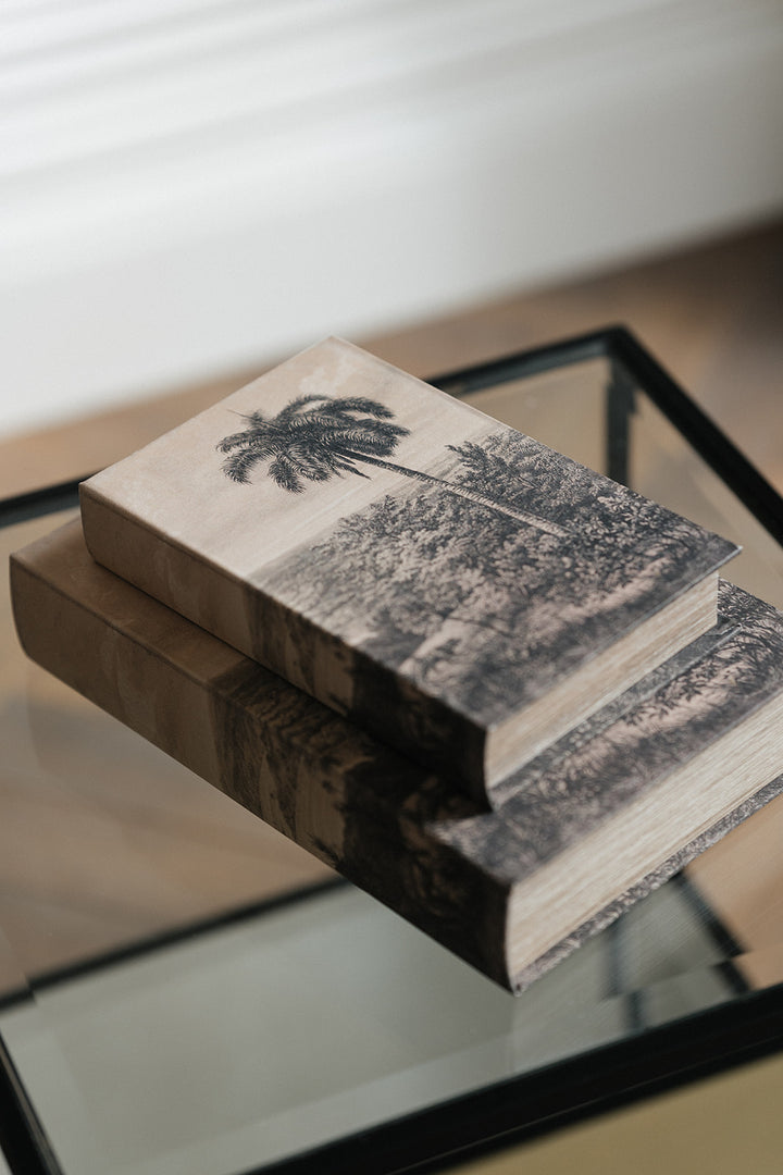 Two decorative books stacked on a glass tabletop, featuring a black-and-white palm tree landscape print on the top book.