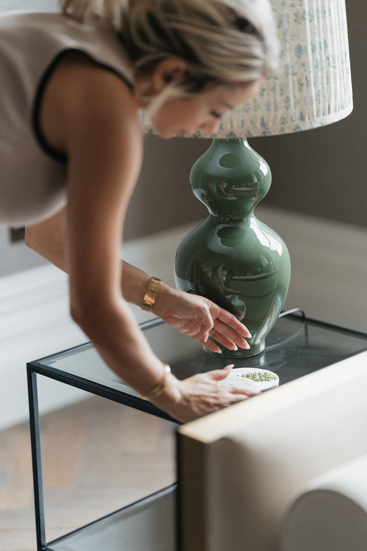 A woman in a beige sleeveless top leans over a glass-top side table, adjusting a glossy green ceramic table lamp with a patterned white shade in a modern living room setting.
