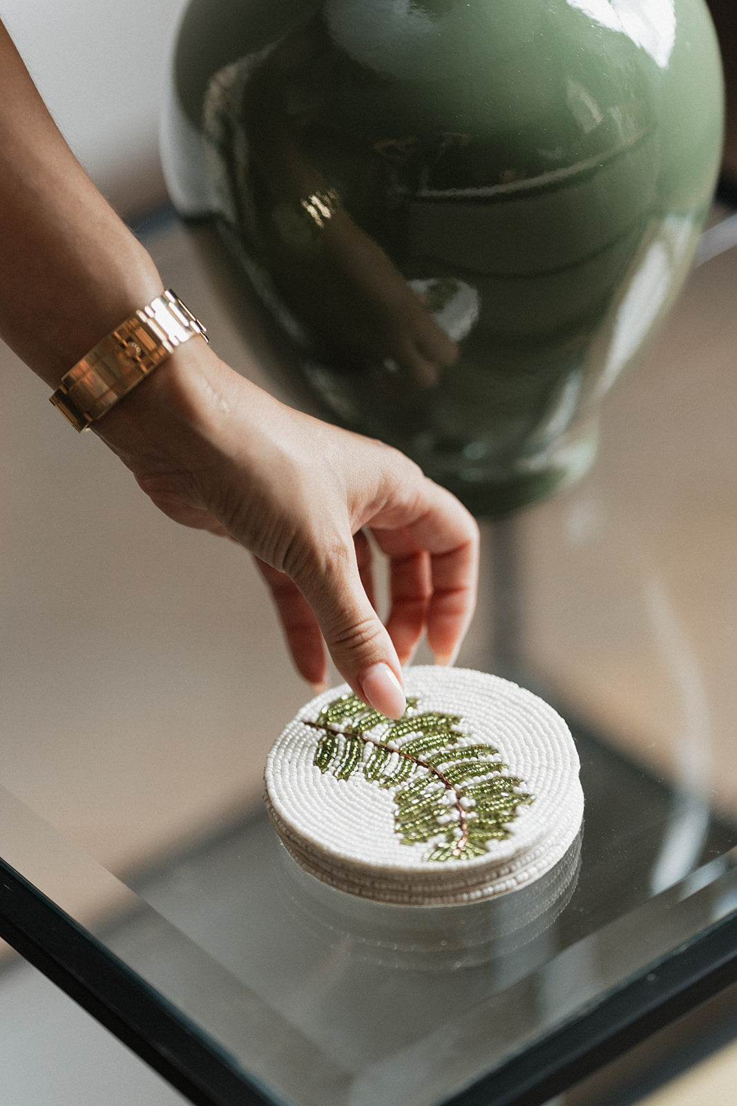 A close-up of a hand wearing a gold bracelet placing a round white woven coaster with a green fern pattern onto a glass table next to a large green ceramic vase.