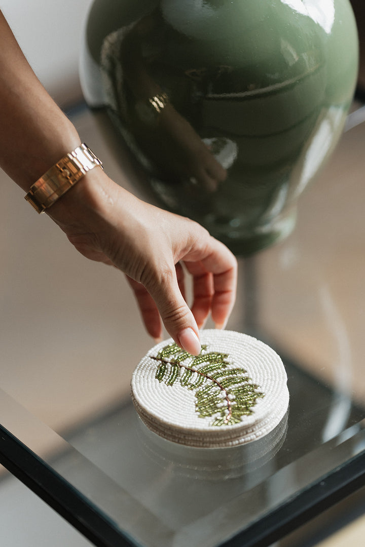 A close-up of a hand wearing a gold bracelet placing a round white woven coaster with a green fern pattern onto a glass table next to a large green ceramic vase.