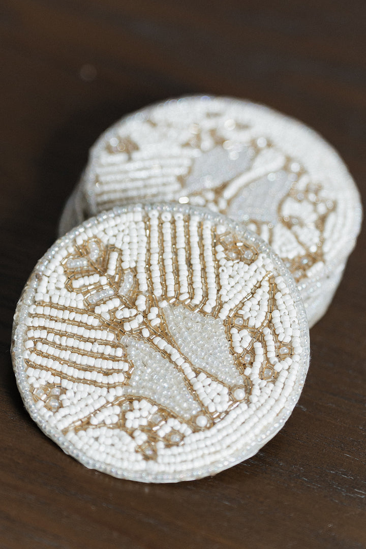 Two round white beaded coasters with gold-toned geometric and floral patterns, stacked on a dark brown wooden surface.