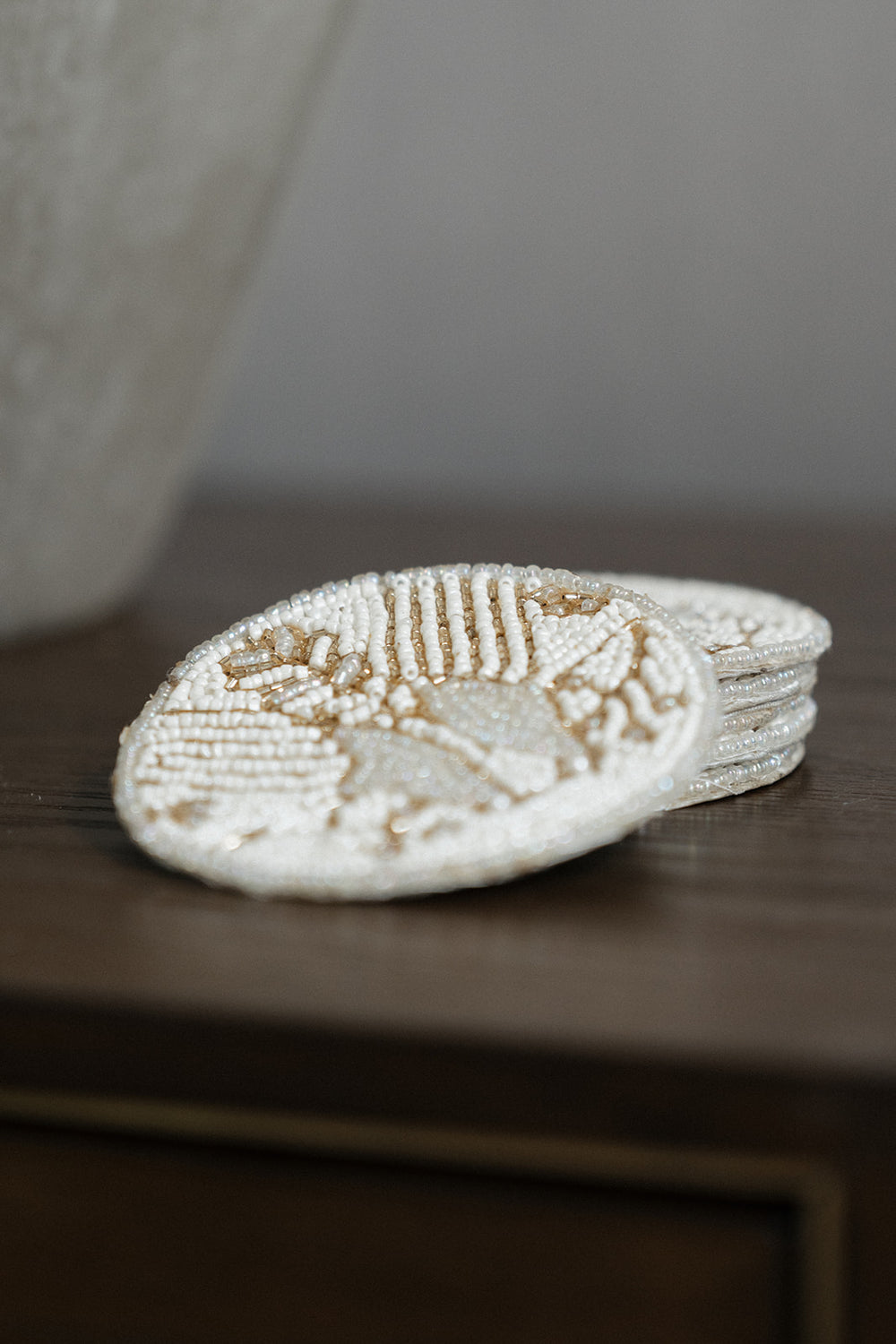 A stack of round, hand-beaded coasters in a cream and light brown color combination, resting on a dark wooden surface.