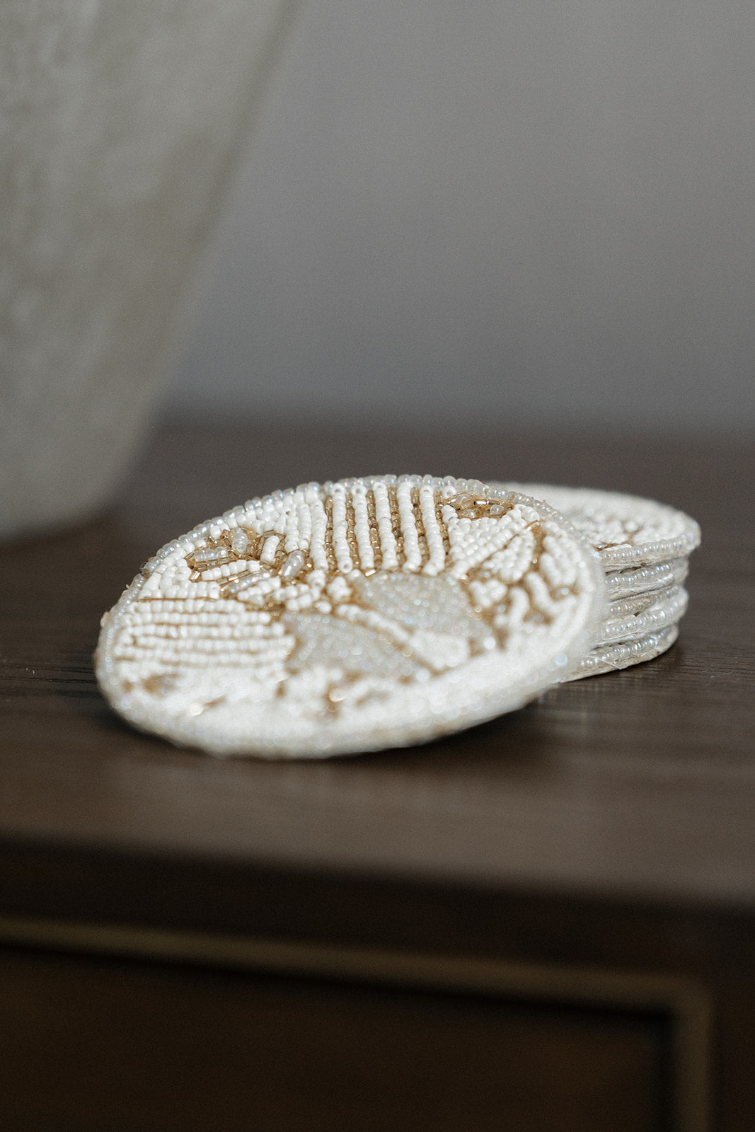 A stack of round, hand-beaded coasters in a cream and light brown color combination, resting on a dark wooden surface.