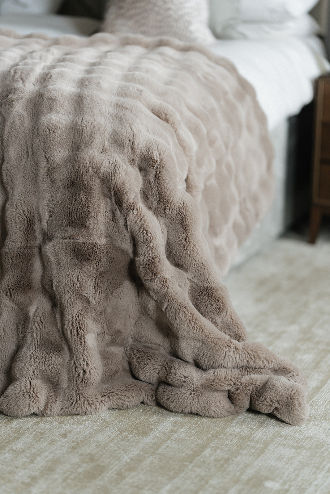 A close-up of a plush, textured throw blanket in a warm taupe-beige color draped over the corner of a bed in a softly lit bedroom.
