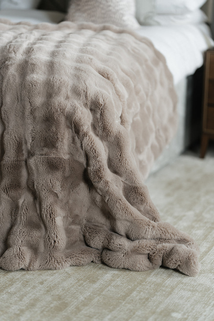 A close-up of a plush, textured throw blanket in a warm taupe-beige color draped over the corner of a bed in a softly lit bedroom.
