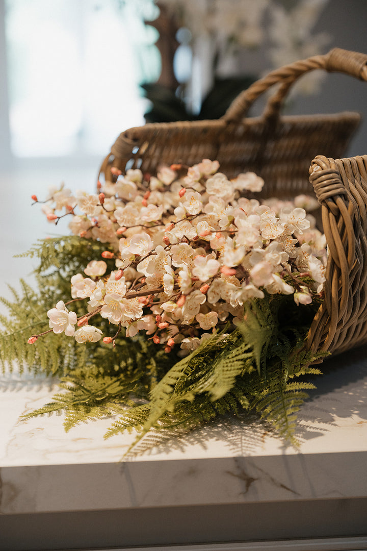 A decorative floral arrangement with pale cream blossoms, green ferns, and small pink buds, placed beside a woven wicker basket on a light marble surface.