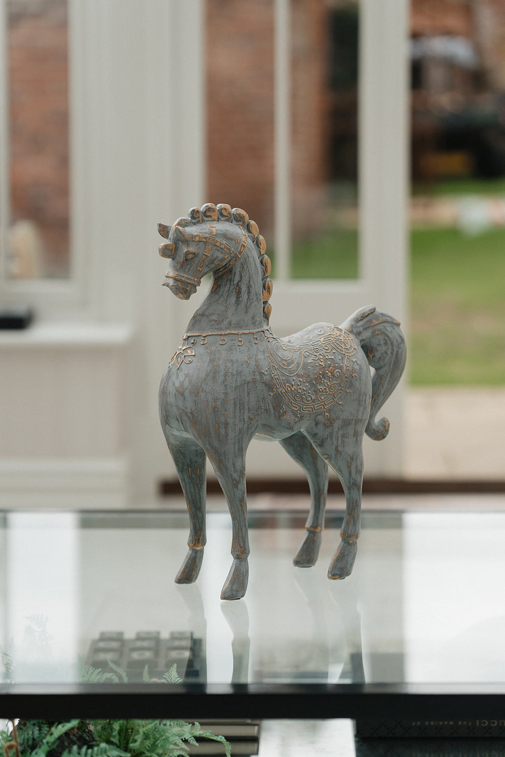 A decorative horse figurine with a distressed gray-blue finish and gold detailing, standing on a reflective glass tabletop in a bright indoor setting.