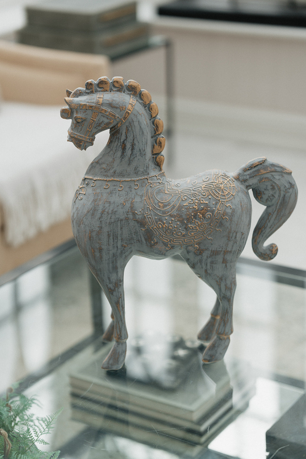 A decorative horse figurine with a distressed gray finish and gold-toned ornamental detailing, displayed on a glass-topped table in a modern interior setting.