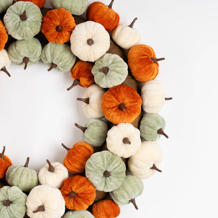 A decorative wreath made of small faux pumpkins in orange, sage green, and off-white, arranged in a circle on a plain white background.