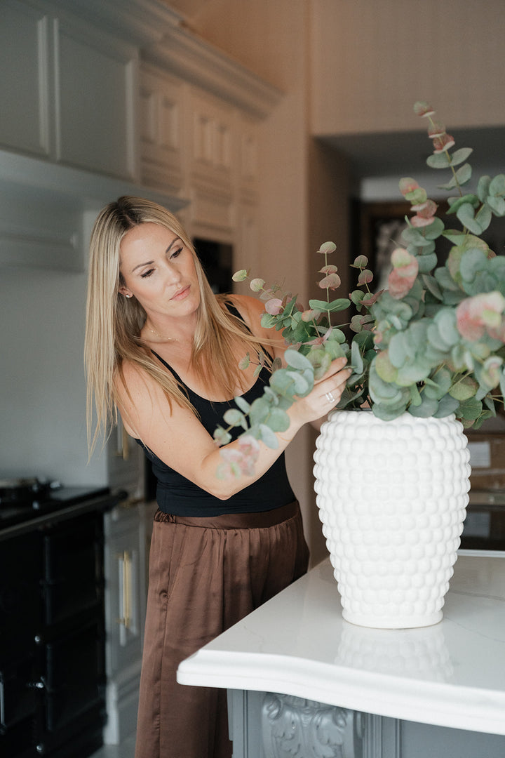 A woman in a kitchen adjusts a large white textured vase filled with eucalyptus branches on a white countertop.
