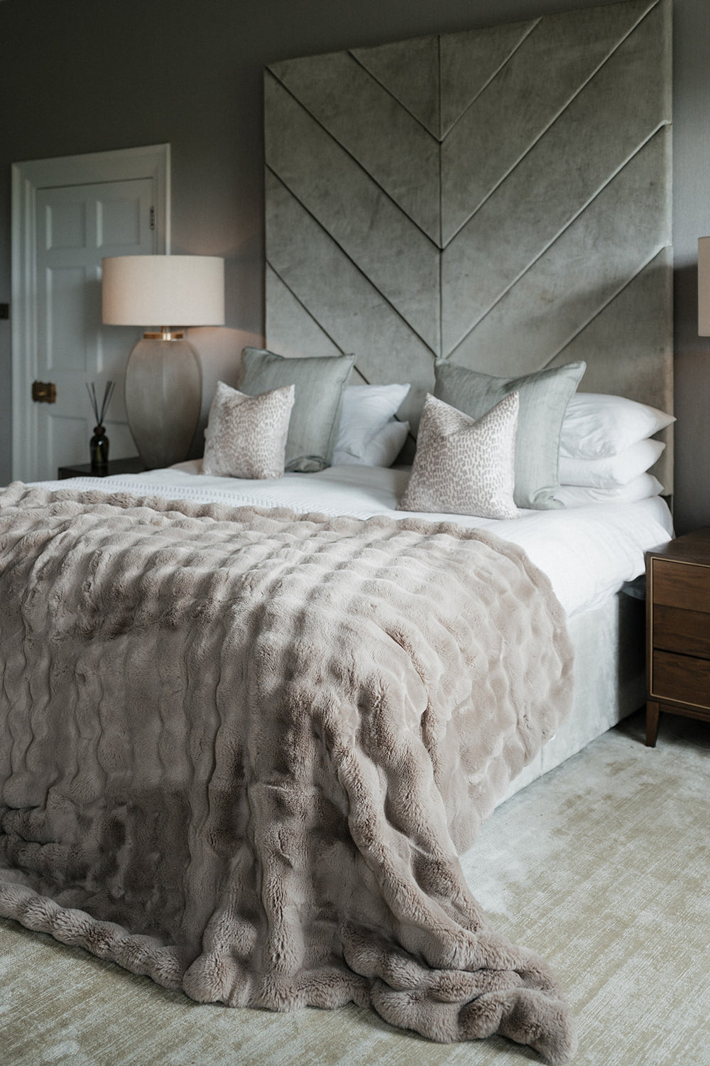 A styled bedroom featuring a bed dressed in white linens, a chunky taupe faux-fur throw blanket, and textured neutral pillows, set against a large gray chevron upholstered headboard.